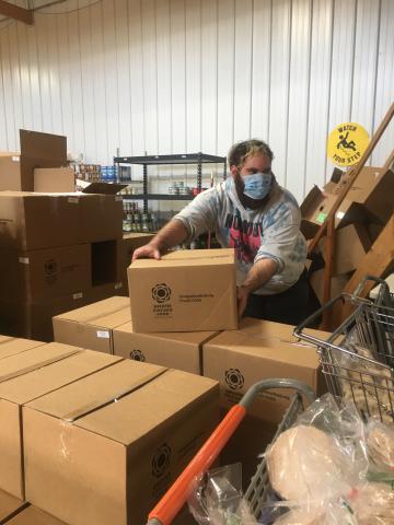blue_cap_food_pantry.jpg A male wearing protective face mask lifts boxes in pantry warehouse setting
