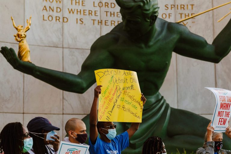 Six protesters (masked for COVID) stand and hold signs in front of the Spirit of Detroit statue in front of Detroit's City Hall
