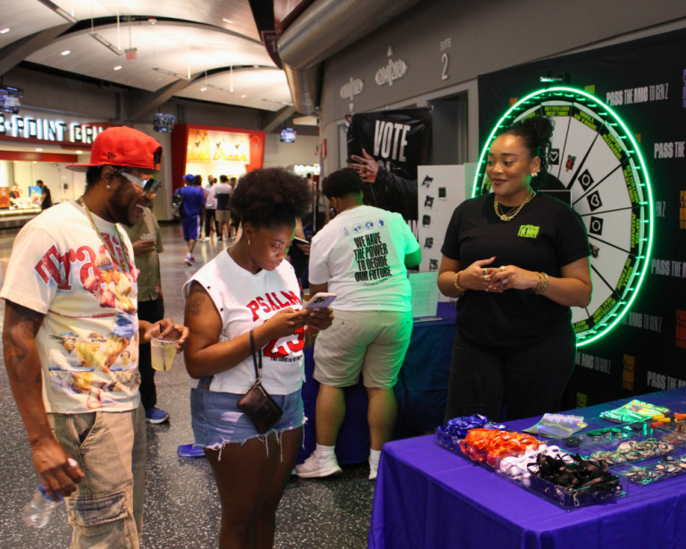 Two people approach a purple table that is covered in giveaway items at a When We All Vote event.