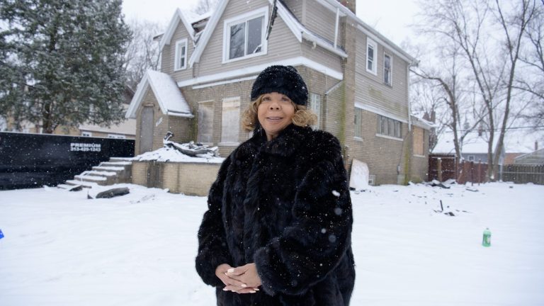 U-Snap-Bac Executive Director Linda Smith, in a winter hat and coat, is standing on a snow-covered lawn in front of a two-story house in Detroit.