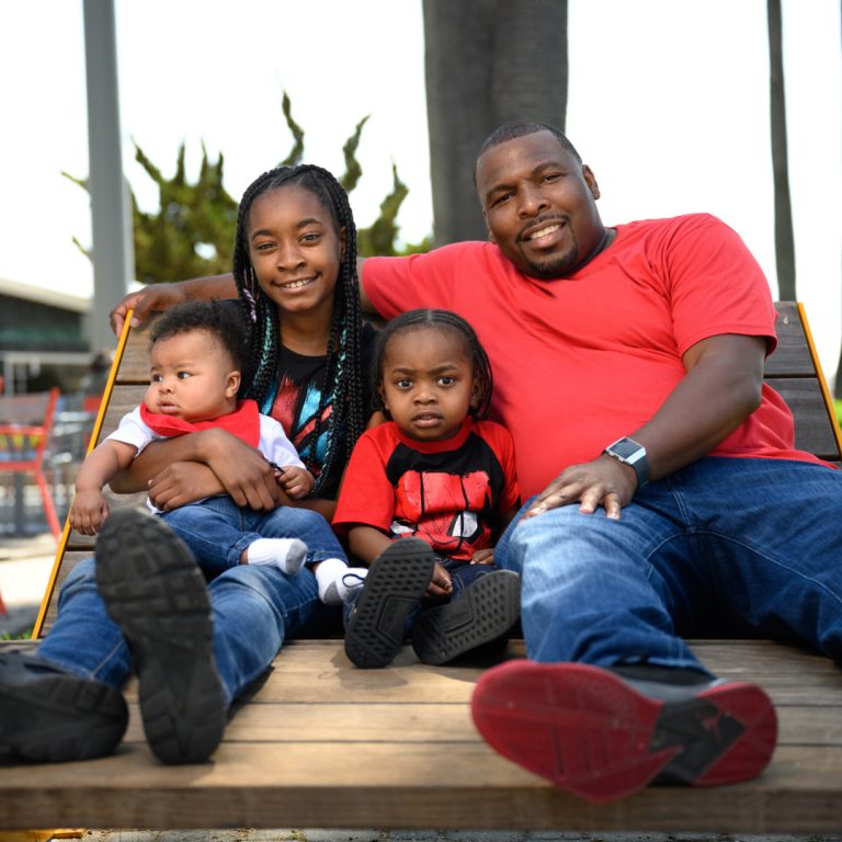 A Black family of a mother and a father and two children sit together and pose for the camera.