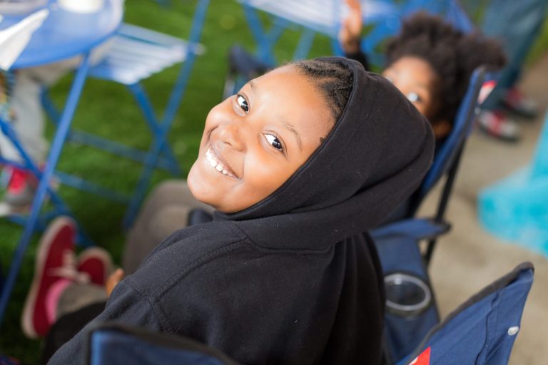 A young Black woman is smiling while sitting on a chair wearing a black sweatshirt.