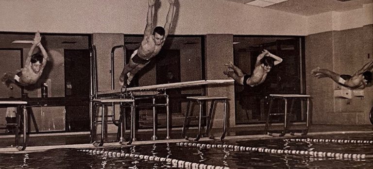 A black and white photo of several swimmers diving into a pool. 