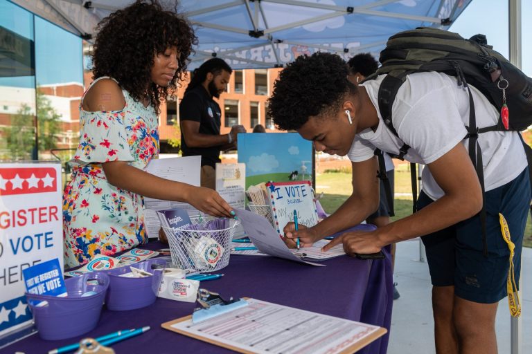 A photo of college students outside at a table with a 'Register to Vote Here' sign, application forms and voting information.