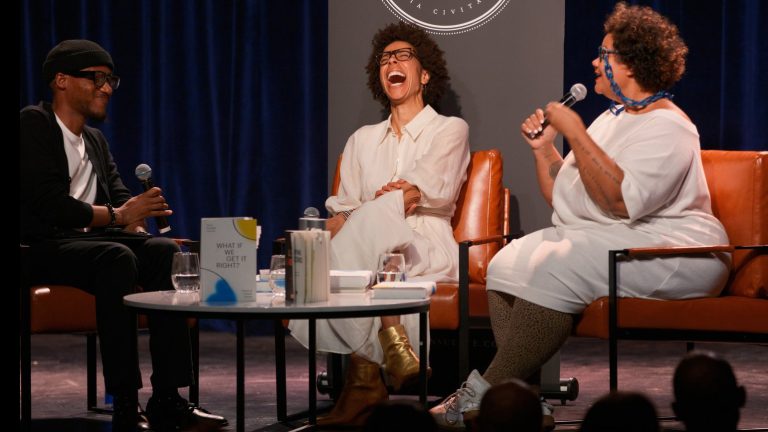 Three people, one Black man and two Black female authors, sit in chairs on a stage holding microphones. One woman is speaking and the other is joyously laughing.