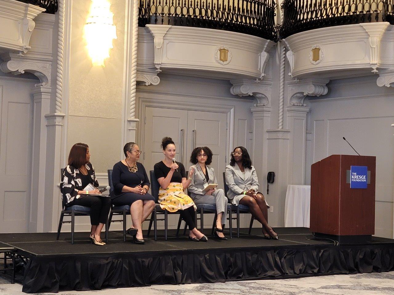 Four women sit on chairs on a stage next to a podium.