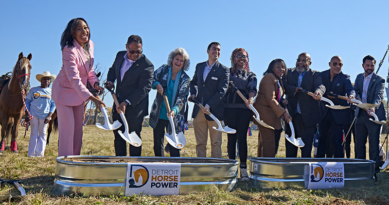 In a field, a row of officials and supporters with shovels turn over dirt in an offical groundbreaking for the Detroit Horse Power equestrian center.