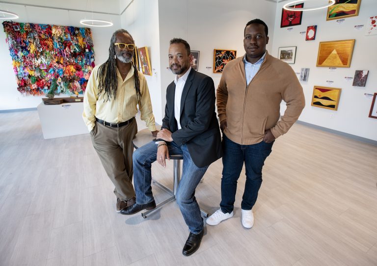 Two men standing and one sitting in the center of the Hannan Center art gallery with multiple art installations on the white walls behind them. 