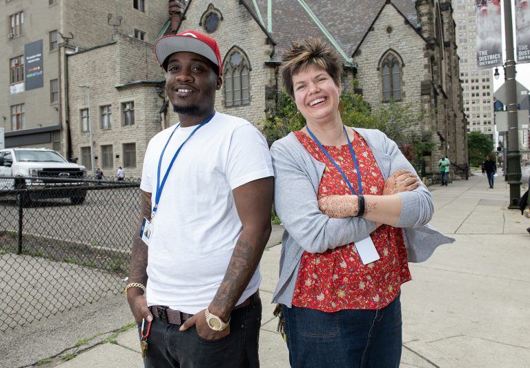 A man in a white T-shirt and red hat and a woman in a red shirt and light blue sweater stand back to back in front of the the Central United Methodist Church in downtown Detroit. 