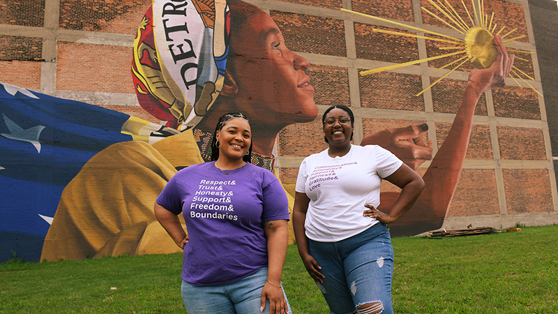 Two people, eleven24 Executive Director Tonjie Reese with a purple shirt that reads “Respect & Trust & Honesty & Support & Freedom & Boundaries” and Program Coordinator Erica Mickens with a white shirt that reads “Communications & Autonomuy & Support & Fairness & Gratitude & Love”, are standing in a field in front of the “Spirit” mural of a woman in a bandana with the word Detroit on it holding a shining orb.