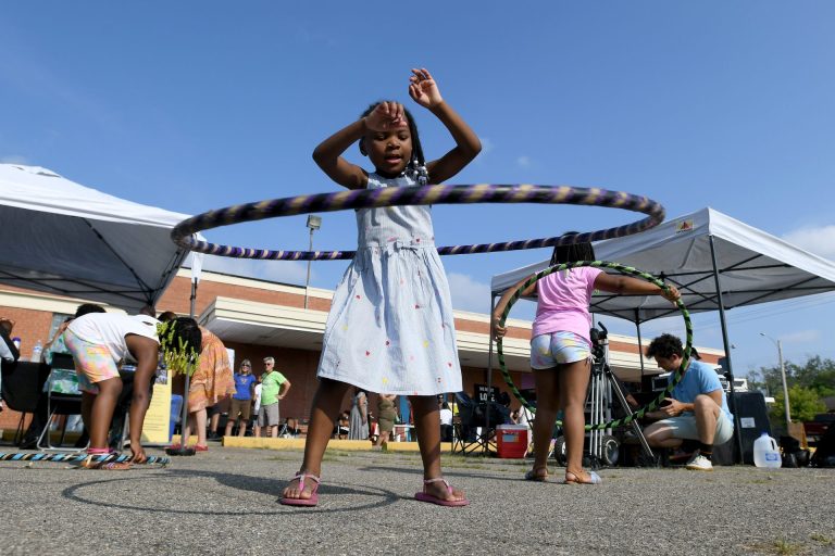 Several children play with hula hoops in a community center parking lot with tables and umbrellas as residents and family members watch during a Sidewalk Detroit Neighborhood Festival.