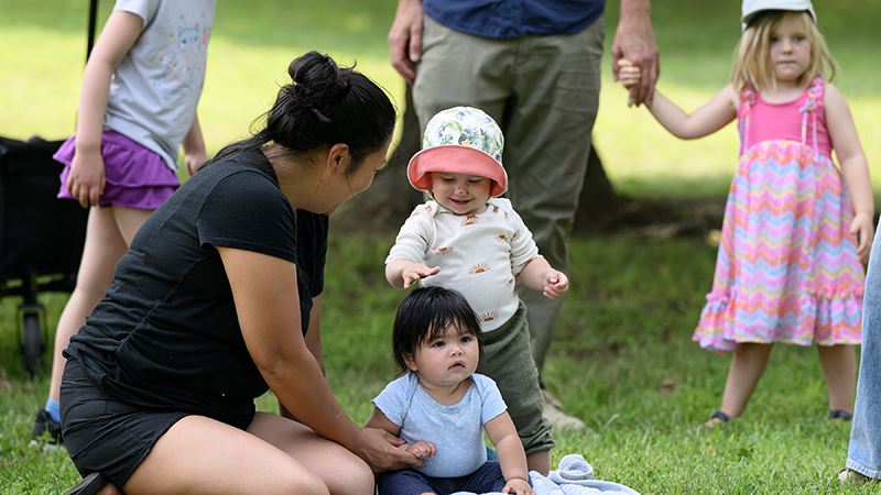 A woman sits on the grass with several children playing in the park.