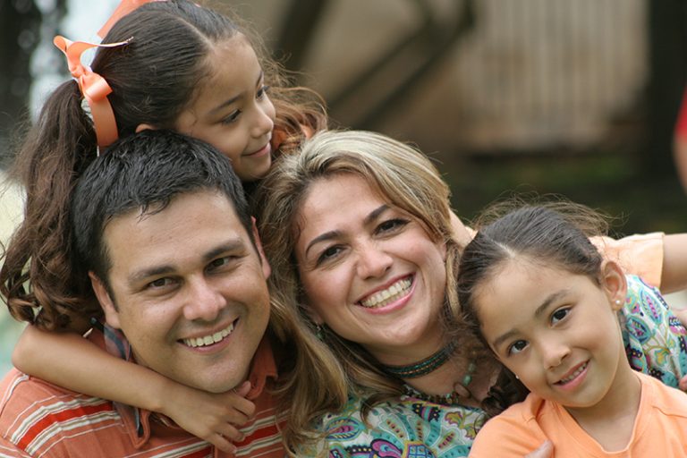 A family of four are seen close together, smiling,and embracing.