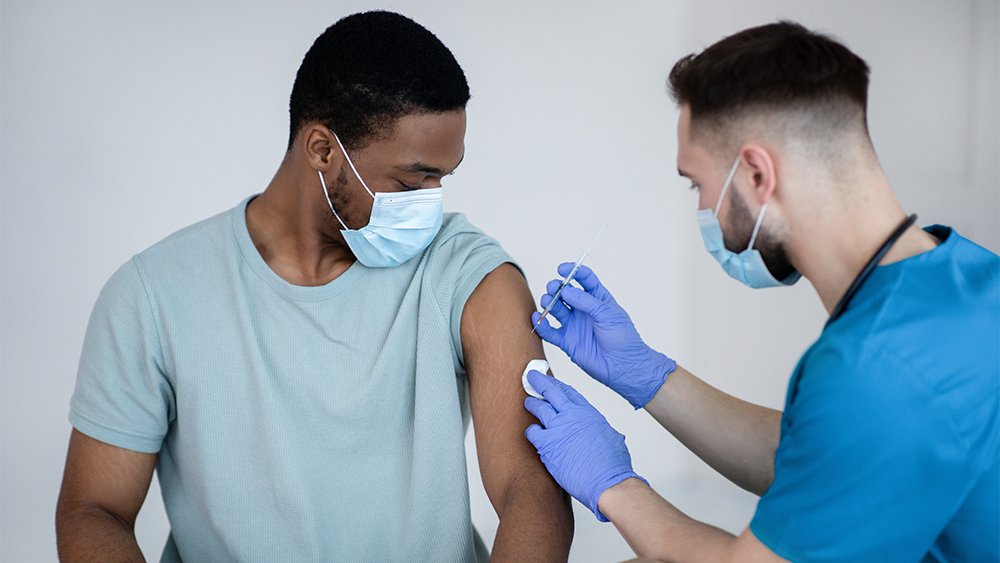 The image is of a man wearing a light green short-sleeved shirt and a mask receiving a shot in his left arm from a man wearing blue medical scrubs, purple medical gloves and a mask.