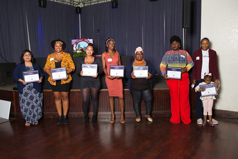 A diverse group of seven people ane child are standing in front of a stage holding certificates from a small business program.