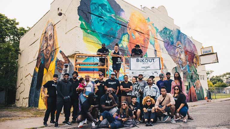 A large group of artists are gathered in front of a colorful mural on a white building with several artists on a lift with the sign BLKOut Walls.