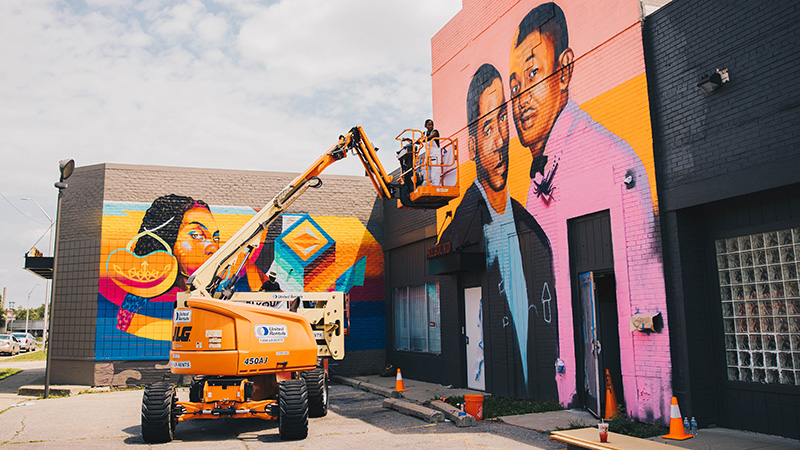 An artist on an orange lift is painting a mural on the side of a wall. 