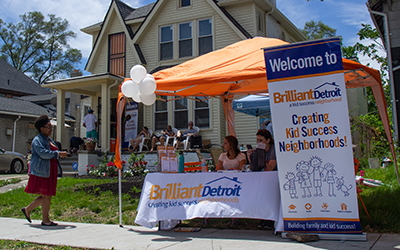 On a sidewalk in front of a house, two people sitting at a table with a Brilliant Detroit banner and an orange canvas above it. 