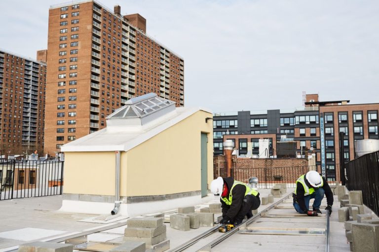Two people in hard hats and safety vests are working on the rooftop of a building in New York CIty. Taller apartment buildings are seen in the background.