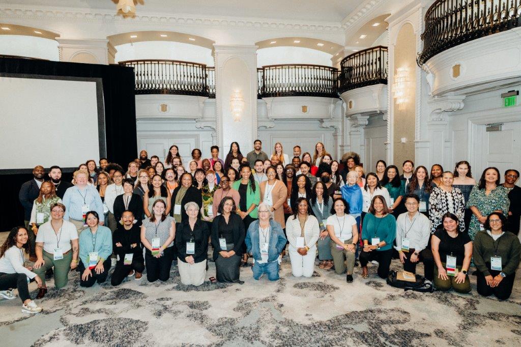 A group of more than 100 people stand in a hotel ballroom to pose for a group photo.