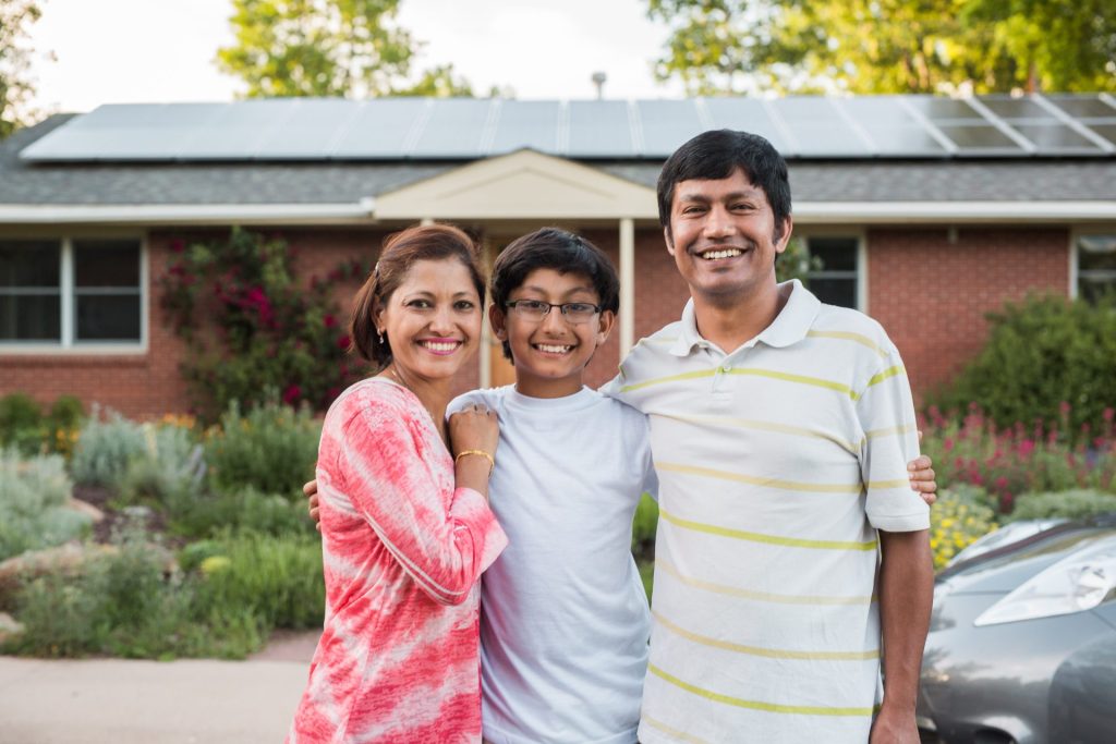 A family of three stand in front of their ranch-style house with solar panels on the roof.