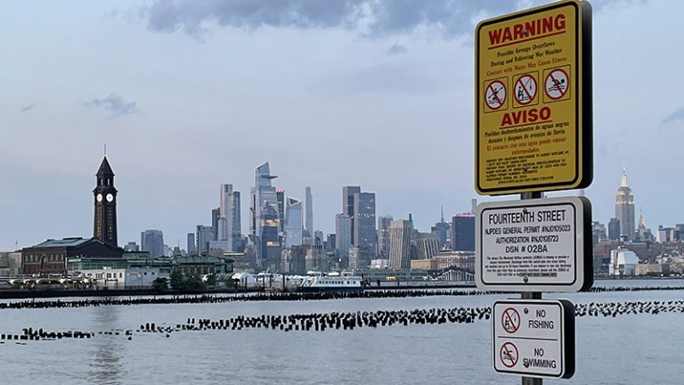 A sewage overflow warning sign on the side of a river with a city skyline in the background