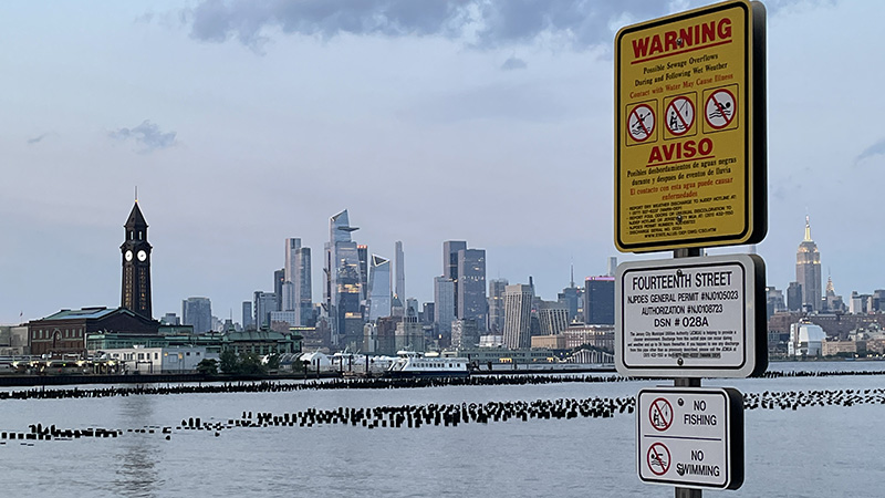 A sewage overflow warning sign on the side of a river with a city skyline in the background