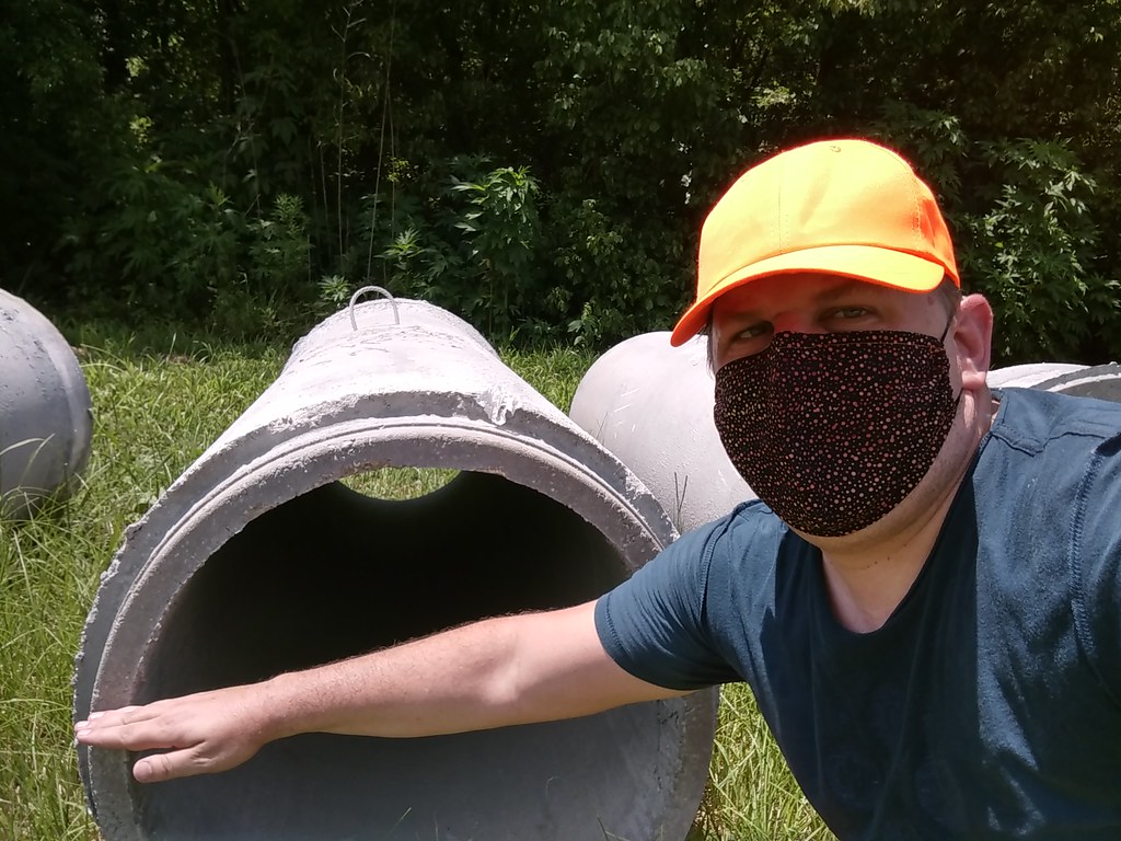 A man in a yellow hat and wearing a black mask has his hand stretched out in front of a large drainage pipe