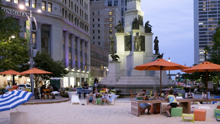 A photo of Campus Martius, a public square in downtown Detroit, with a large monument with statutes of soldiers in the center and surrounded by sand and people sitting at umbrella-covered tables. The Detroit skyline is seen in the background.