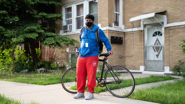 Roosevelt High School student Nathaniel Martinez, in a blue jacket and red pants, stands with his bikeon the sidewalk in front of his house, a beige brick building with a white door.