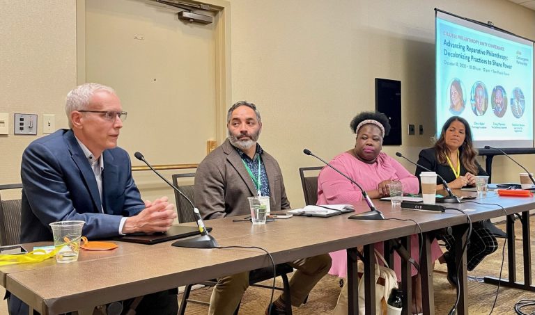 A panel of four people sit at a table with a screen to the right that reads: Change Philanthropy Unity Conference | Advancing Reparative Philanthropy: Decolonizing Practices to Share Power