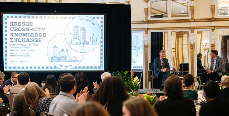 Two men in suits sit on a stage with a large screen that reads Kresge Cross-City Knowledge exchange.