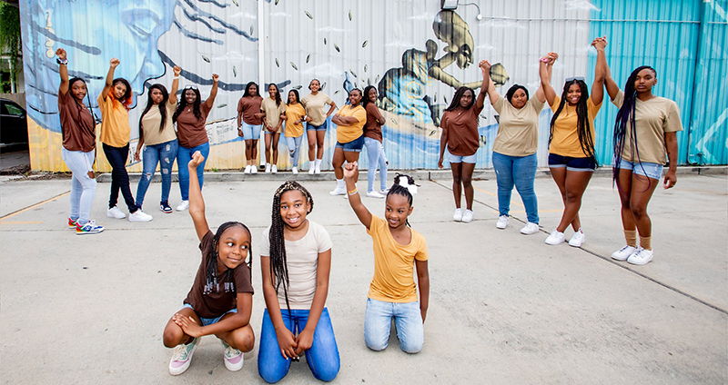 A group photo of young Black girls with their arms raised in an empowering pose, standing outside in front of a mural.