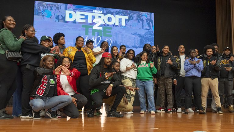 A large group of students and fraternity members are gathered on a stage with a sign behind them that reads From Detroit 2 the Yard.