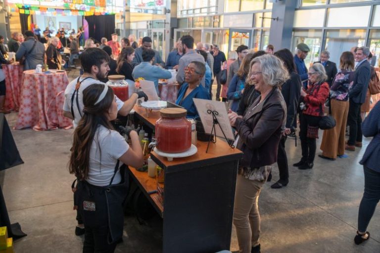 A large group of people at the MexicanTown CDC Mercado in southwest Detroit at the Eminent Artist monograph celebration with food, drinks and colorful decorations.