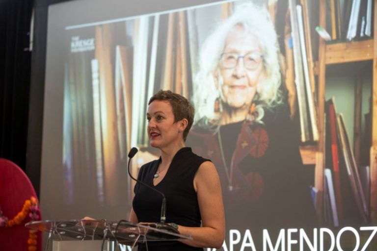 Kresge Arts in Detroit Deputy Director Katie McGowan speaks at a lectern with an image of Kresge Eminent Artist Nora Chapa Mendoza on the screen behind her.