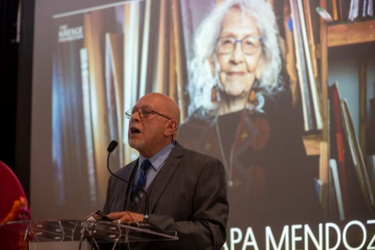 Ozzie Rivera, a southwest Detroiter and community activist, speaks at a lectern with an image of Kresge Eminent Artist Nora Chapa Mendoza on the screen behind her.