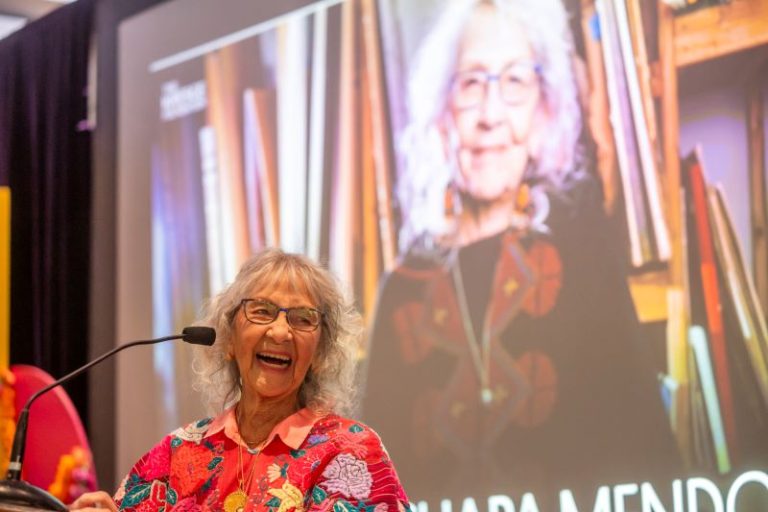 Nora Chap Mendoza speaks at a lectern with an image of herself behind her on a screen.