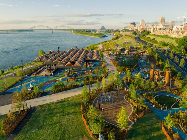 An aerial drone view of the renovated Tom Lee Park on the riverfront in Memphis with a sunset canopy, trails, fitness area, playground. A bridge spanning the river and the downtown skyline is seen in the background.