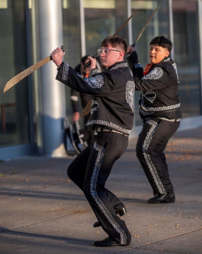 A performance by the Milagro Compañía de Ballet Folklórico with several men in black jacket and pants trimmed in white and holding swords.