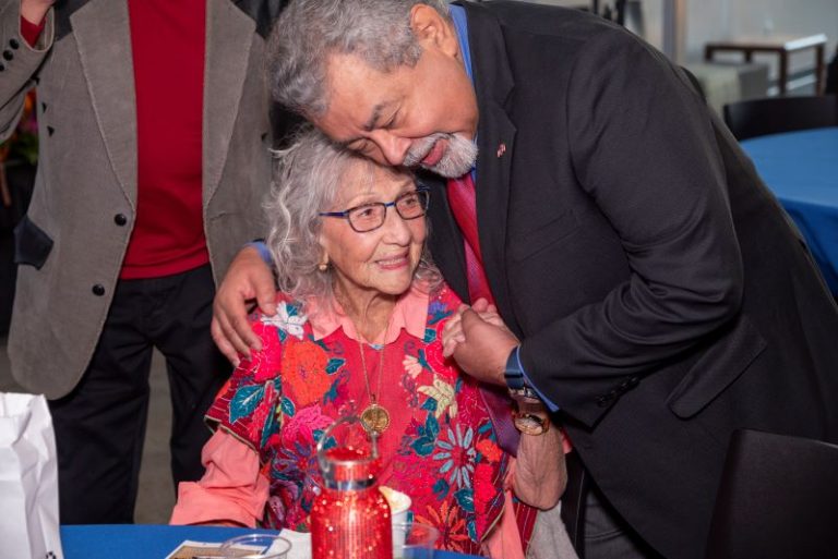 Nora Chapa Mendoza in a colorful red floral outfit gets a hug from a friend