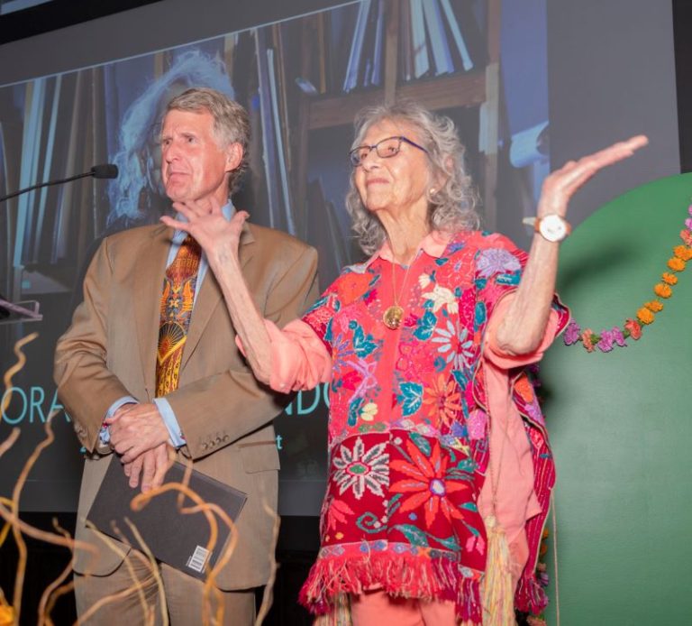 Kresge President Rip Rapson, in a tan suit and holding the Eminent Artist monograph, stands next to Nora Chapa Mendoza in a floral-patterned red outfit as she raises her hands in the air.
