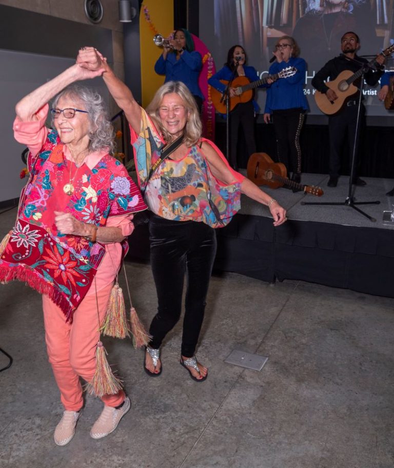 Nora Chapa Mendoza, 92, in a floral-patterned red outfit dances with another woman in front of a mariachi band.