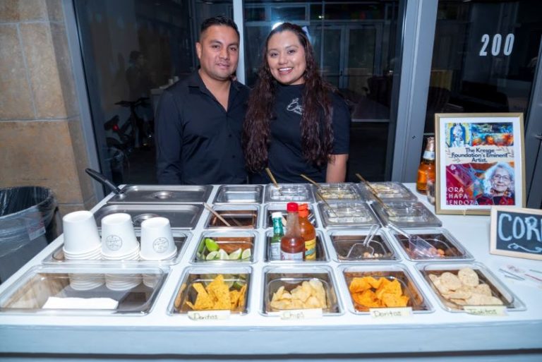 Two food preparers behind a corn salsa bar.