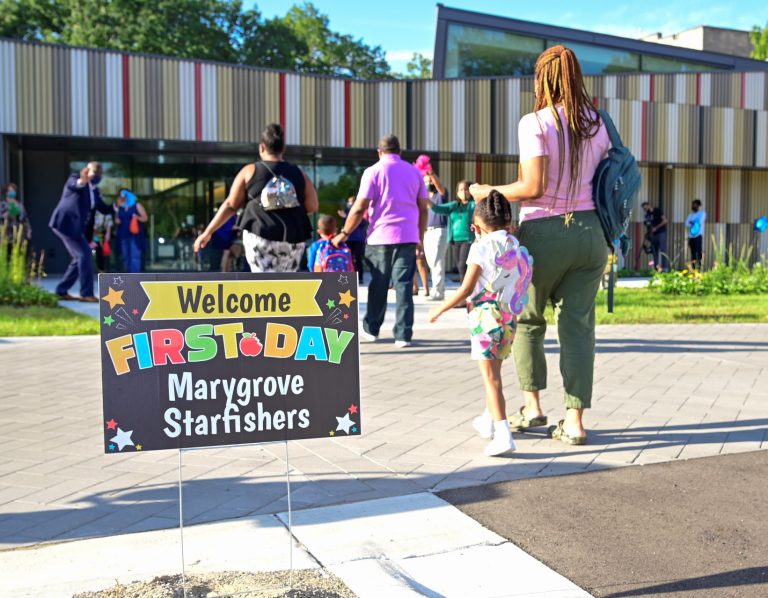Lawn sign reads "Welcome First Day Marygrove Starfishers." Parents and children walk by to childcare center on sunny day.
