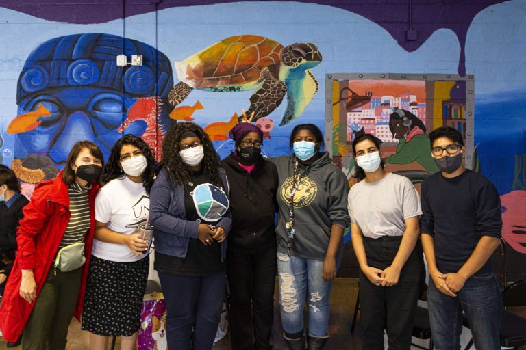 Seven youths or young adults pose in front of a brightly painted wall mural which includes a large turtle, fish and what looks to be a sculpted head.