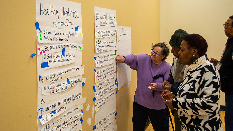 Several housing residents are gathered for a listening session as they cluster around poster boards with comments. 