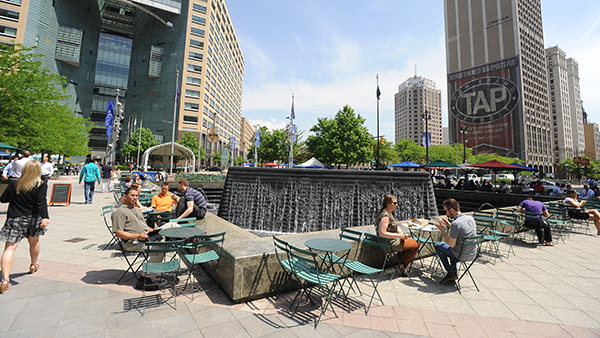 People are sitting at several sets of table and cbhairs in front of a water fountain in the middle of the city square called Campus Martius Park.