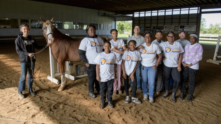 A group photo of people in white Detroit Horse Power T-shirts pose together in a stable with a horse.