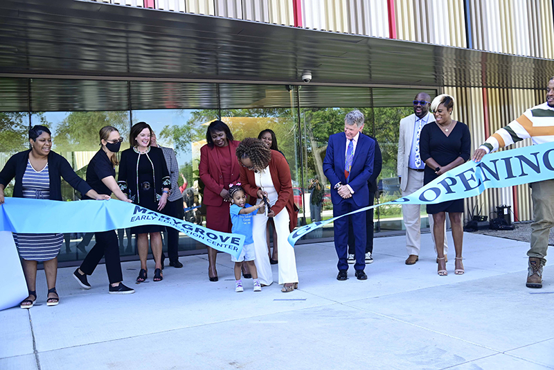 The principal of a school and a young student cut the ribbon that says Marygrove Early Education Center Grand Opening with a row of officials behind them in front of the new school.